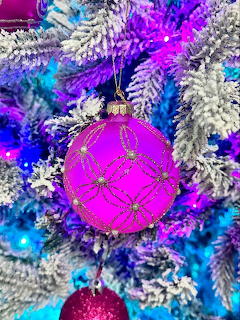 Pink glitter ornament with gold patterns hanging on a flocked Christmas tree with blue and purple lights