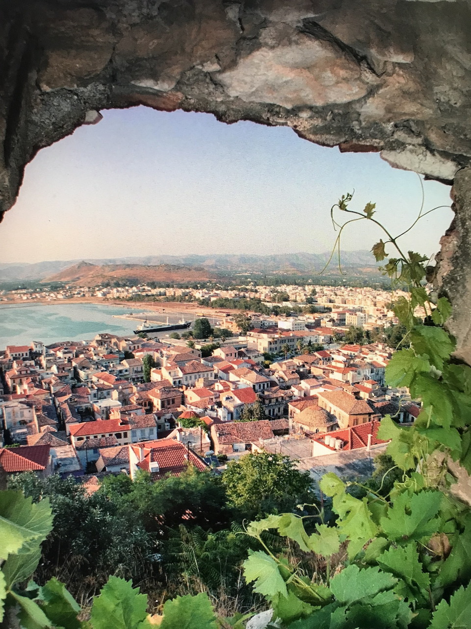 Coastal town with red roofs viewed through rocky cave opening with vines, sea visible in background
