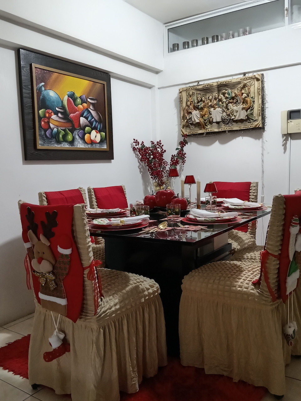 Dining room with Christmas-decorated chairs, red accents, set table, and framed paintings on the wall.