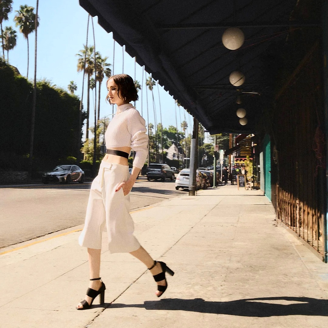 Woman in a white outfit and black heels walking on a sunny city sidewalk
