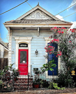 Small white house with red door, black shutters, brick path, and garden plants