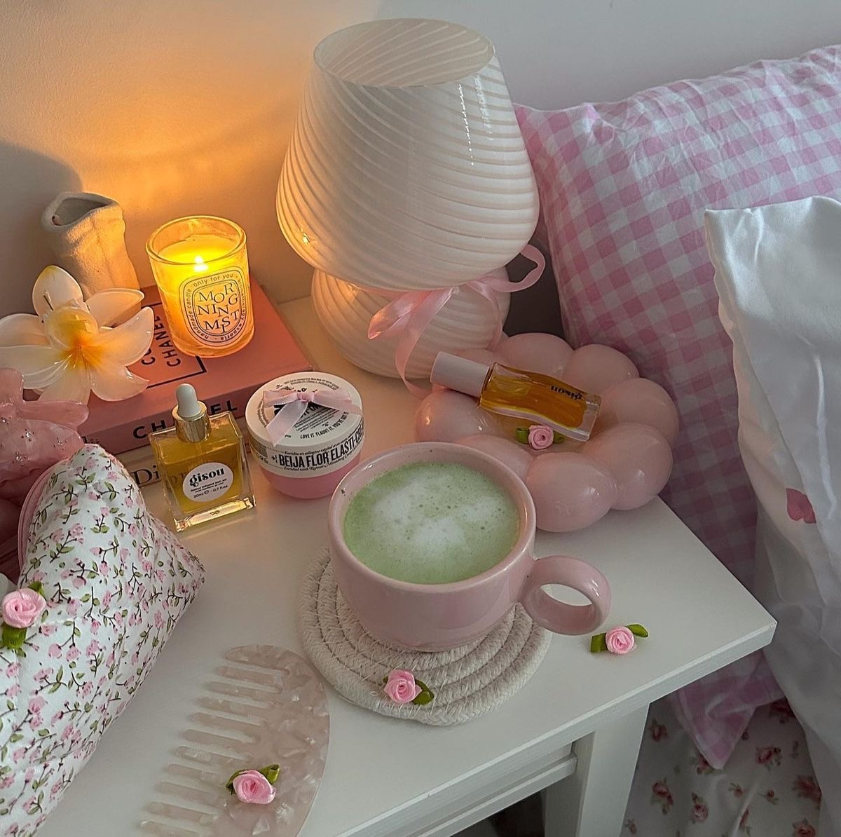 Pastel bedside table with candle, pink lamp, matcha drink, perfume, and floral decor beside a bed