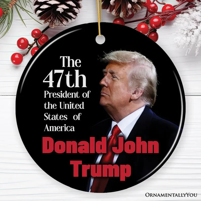 Black Christmas ornament with Donald Trump portrait and printed text, surrounded by pinecones and red berries