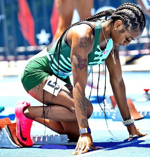 Female sprinter in green uniform positioned in starting blocks on blue track preparing for race start