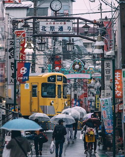 Rainy Japanese shopping street with yellow train and pedestrians carrying umbrellas