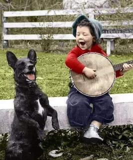Happy child playing banjo with a black and white dog sitting up beside them outdoors