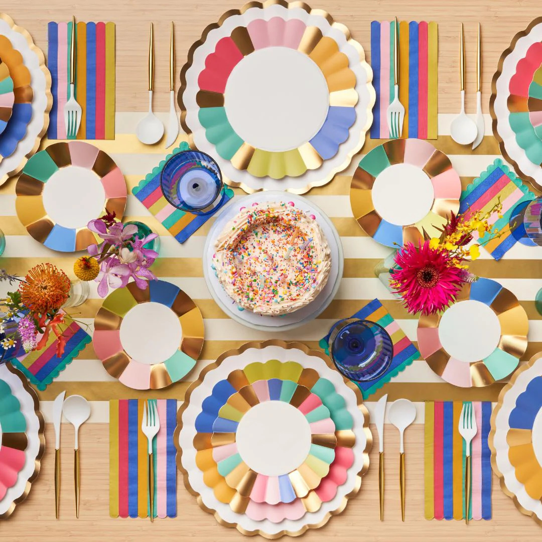 Overhead view of colorful birthday party table with rainbow plates, flowers, and cake