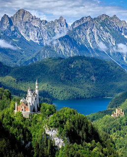Neuschwanstein Castle on a forested hill with a blue lake and Alpine mountains in the background