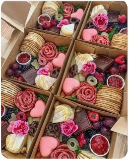 Multiple charcuterie boxes with crackers, salami roses, heart cookies, grapes, berries, cheese, and flowers