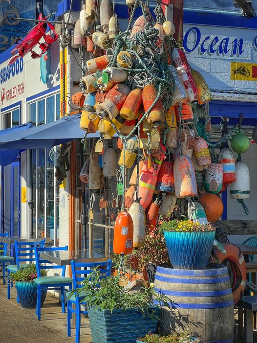 Cluster of orange, yellow and multicolored fishing buoys hanging on weathered post with blue striped building behind