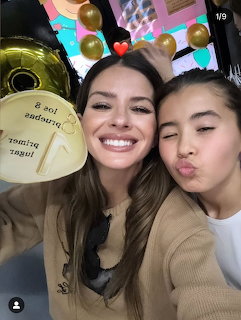 Two smiling women taking a selfie with gold balloons and colorful party decorations behind them.