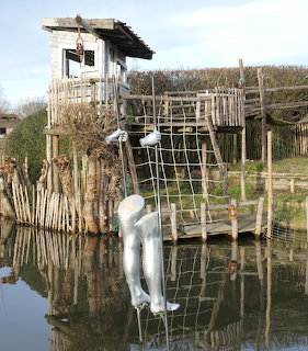 Mannequin hanging on net attached to wooden stilt hut over water in outdoor art installation