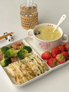 White tray with porridge, strawberries, broccoli, and sliced rolls beside a glass drink