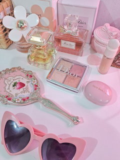Pink desk flatlay with heart-shaped sunglasses, perfume bottles, makeup palette, and vintage hand mirror