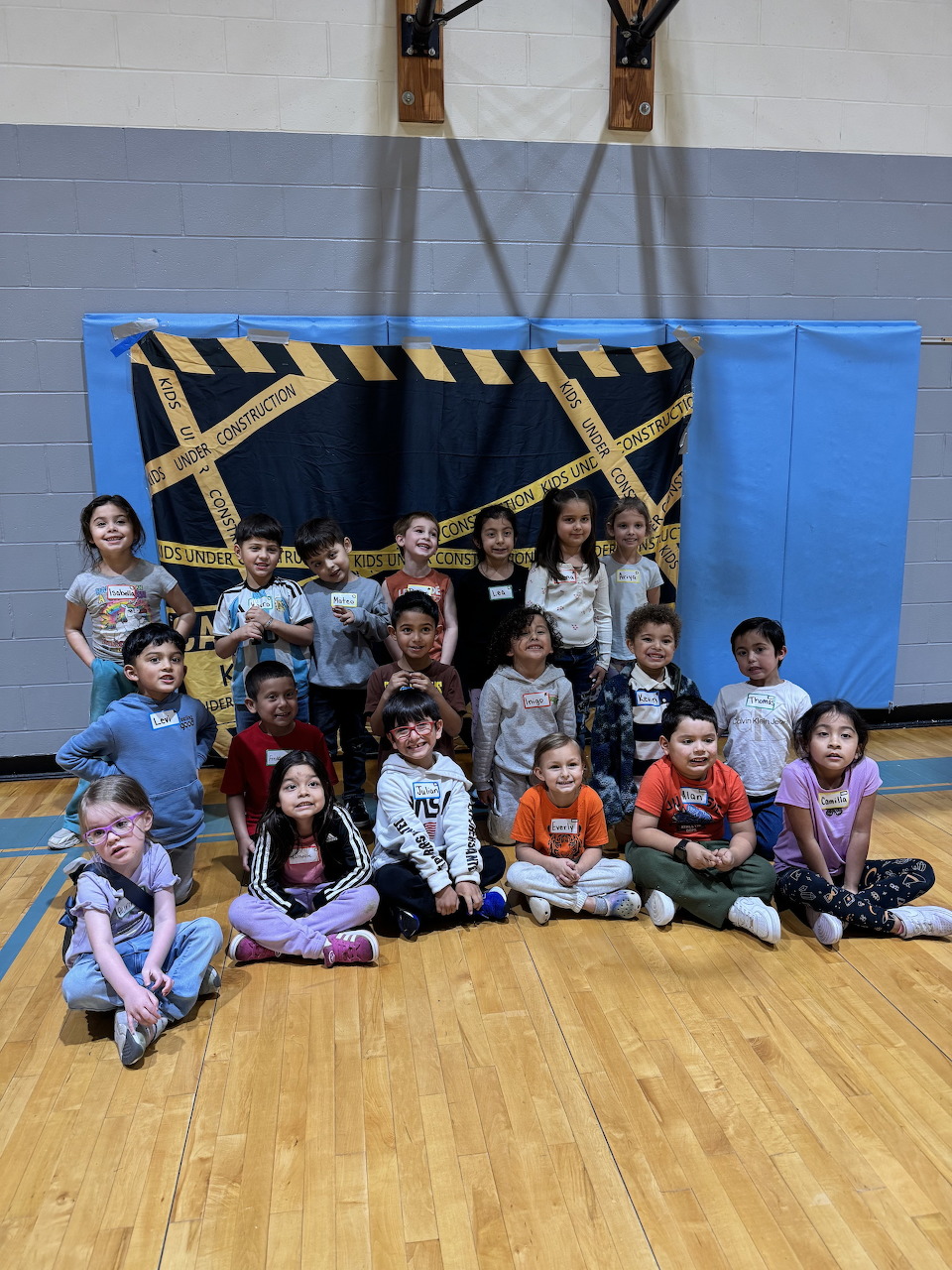 Group of kindergarten children smiling and posing in a school gym in front of a blue and yellow backdrop.