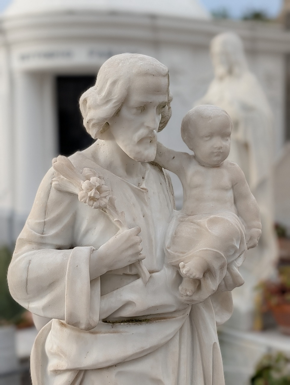 White marble statue of a woman holding a child with blurred mausoleums behind