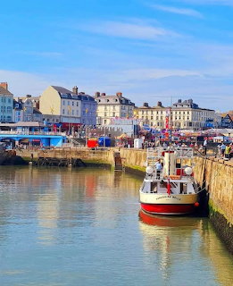 Harbor with boats moored at stone quay, colorful buildings line waterfront under blue sky