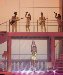 Female performer in yellow sparkly outfit on pink stage with musicians on elevated platform above