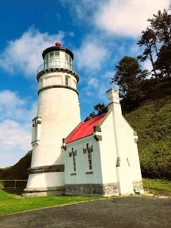 White coastal lighthouse with red roof beside keeper's house on grassy hillside with trees and blue sky
