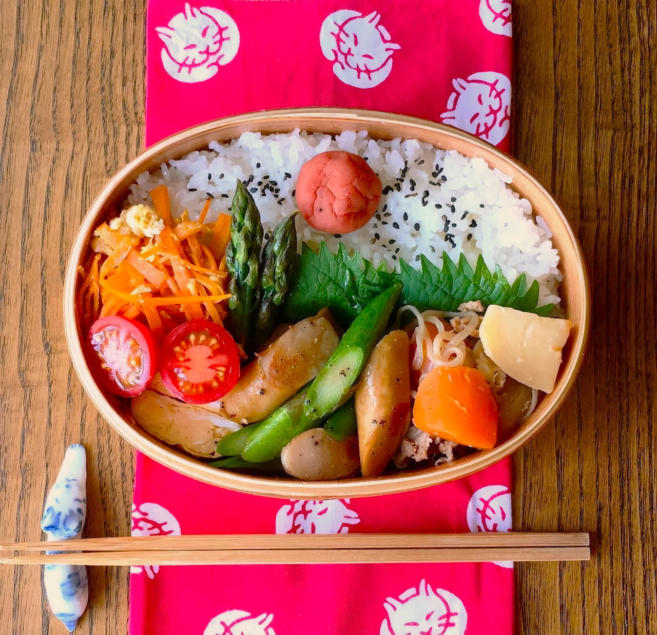 Top-down view of a Japanese bento box with rice, vegetables, sausage, and chopsticks on a wooden table