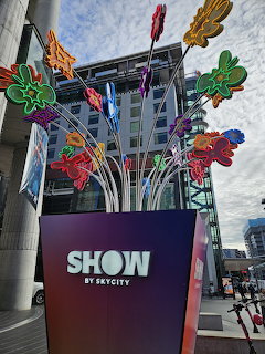 Colorful flower-shaped art installation on metal stems in front of a modern building in Auckland