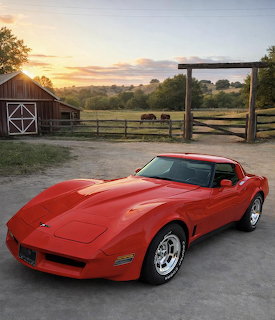 Red C3 Corvette parked on a farm driveway at sunset with a barn and horses behind it