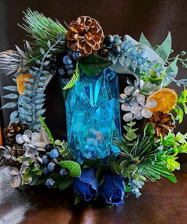 Festive wreath with blue glass ornament, pine cones, evergreen branches, and blue flowers on dark background