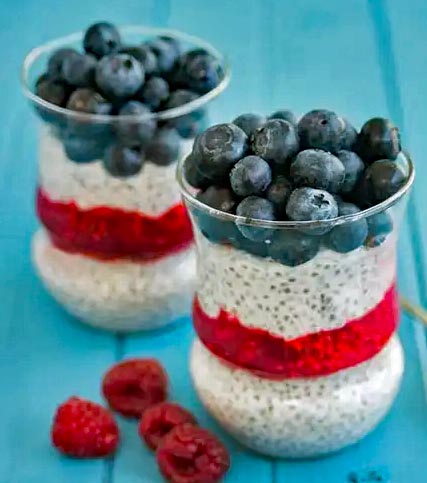 Two glass jars of layered chia pudding with blueberries, raspberries, and cream on turquoise surface