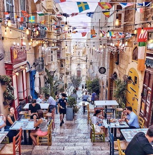 Narrow cobblestone street in Valletta with outdoor cafe seating, historic buildings, and colorful flags strung overhead