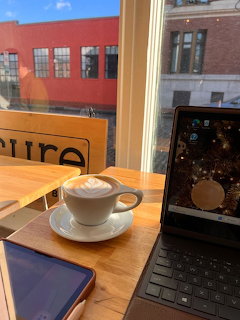 Cappuccino cup on wooden table with laptop and tablet, brick buildings visible through window