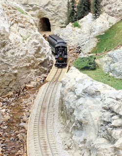 Model train on curved track approaching tunnel entrance in rocky landscape with grass patches