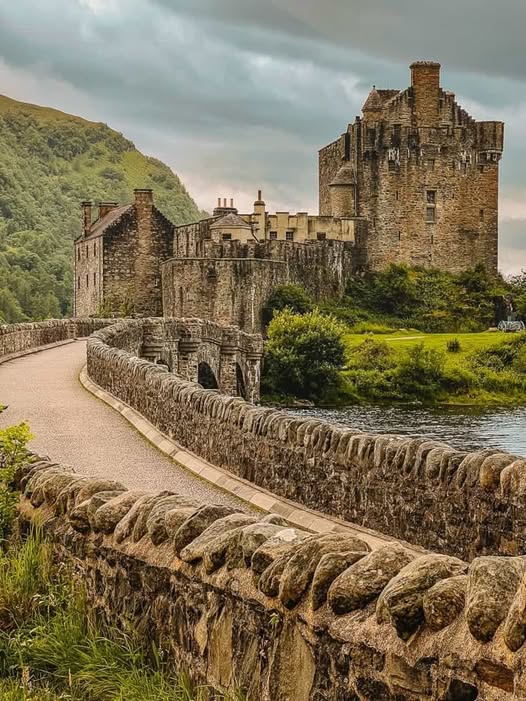 Eilean Donan Castle seen from stone bridge with green hills and cloudy sky in Scotland