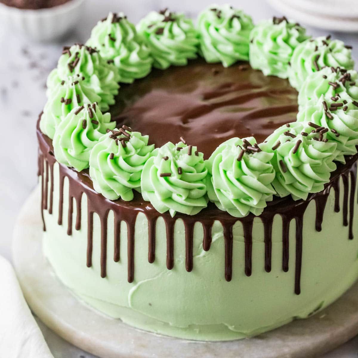 Round mint chocolate cake with green frosting, chocolate drip, and piped mint rosettes with chocolate shavings