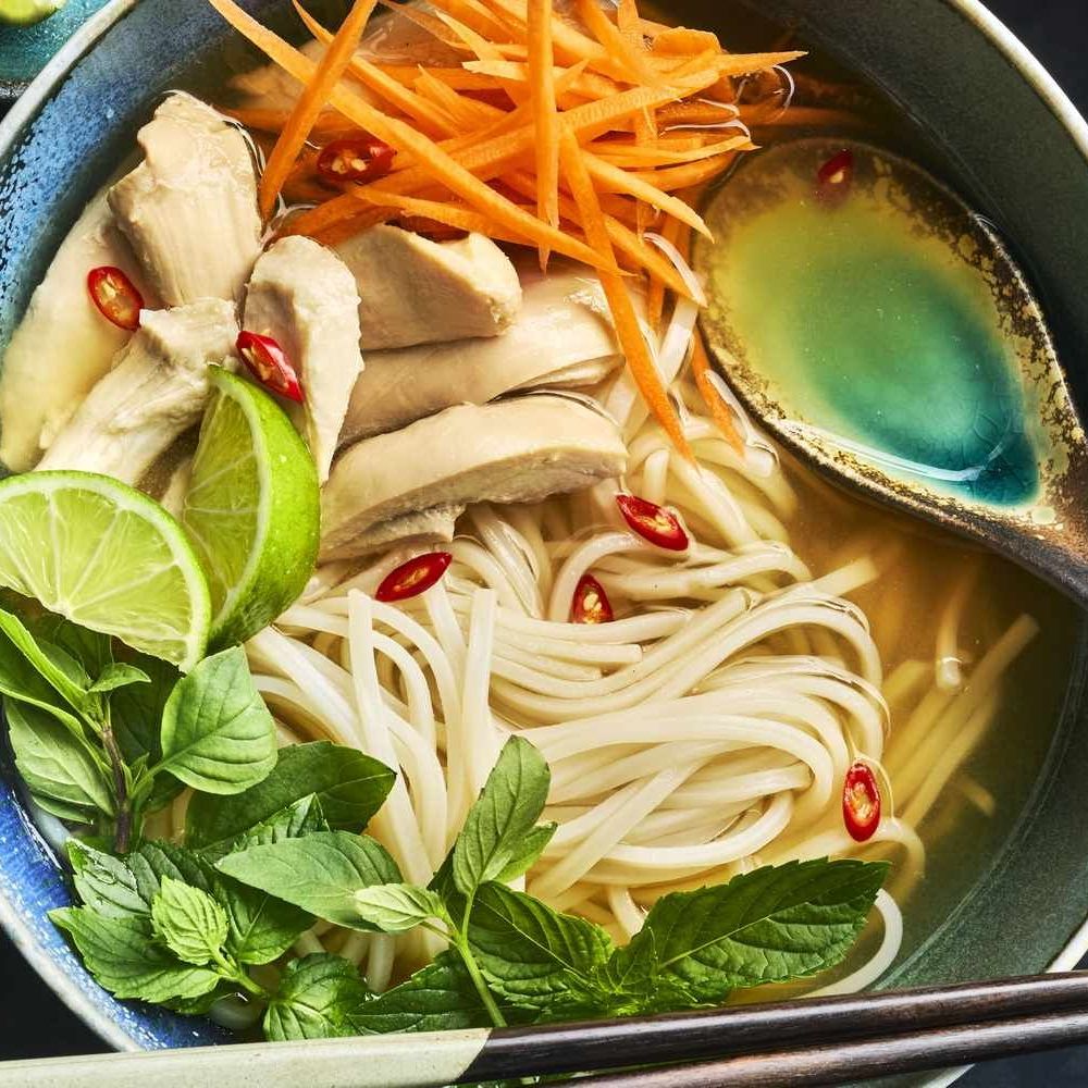 Bowl of pho soup with noodles, chicken, herbs, lime, carrots and chili peppers in broth