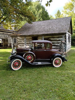 Dark maroon vintage Ford Model A car parked on grass in front of a log cabin with trees behind