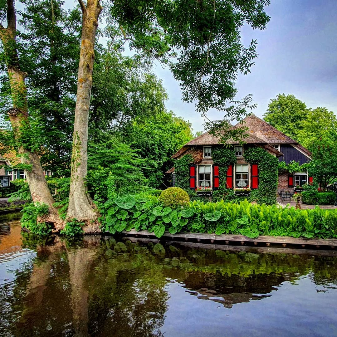 Red farmhouse with thatched roof beside calm canal surrounded by trees in Giethoorn village