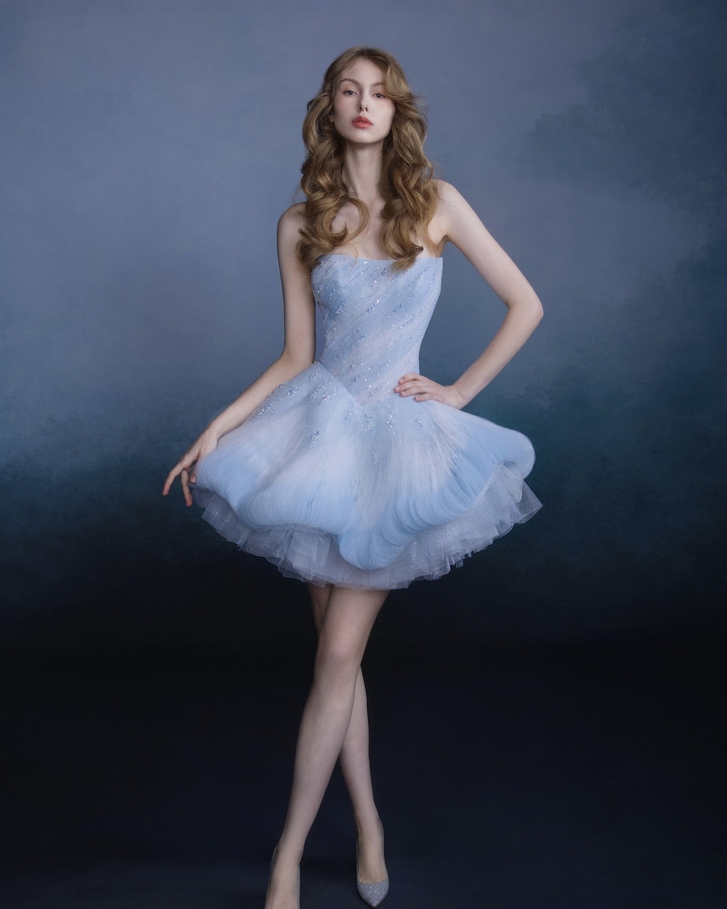 Woman in light blue tulle dress with layered skirt posing against blue studio background