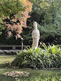 Stone statue of a robed figure standing by a pond surrounded by green plants and autumn trees