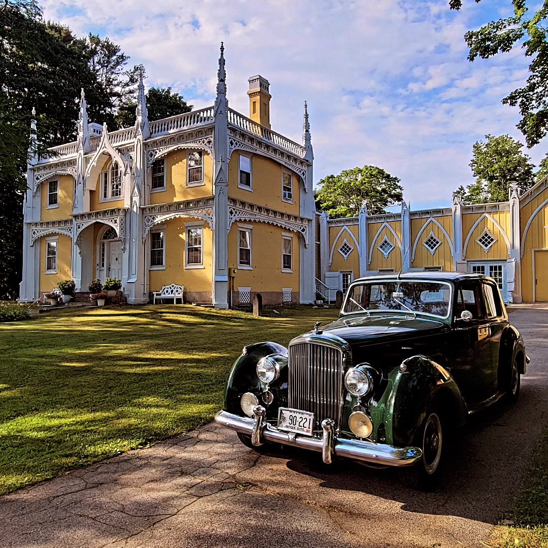 Yellow Gothic Revival mansion with white decorative trim and vintage black car in front on sunny day