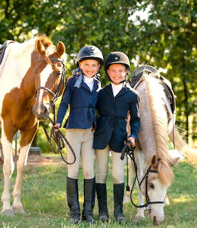 Two young girls in riding gear standing with their horses outdoors on green grass