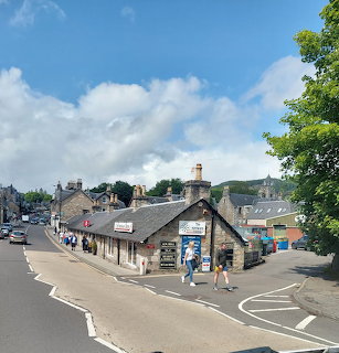 Stone buildings along a village street with pedestrians, trees, and cloudy blue sky
