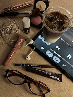 Flatlay of cosmetics, eyeglasses, earphones, iced coffee drink, and a tablet on a wooden surface