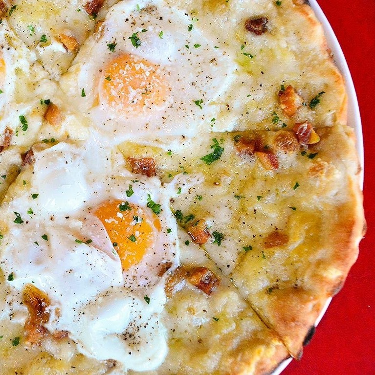 Flatbread pizza topped with three fried eggs, melted cheese, herbs, and bacon on red background