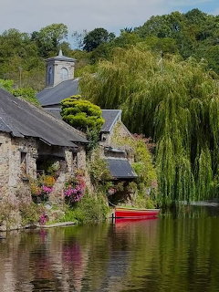 Stone buildings and bell tower beside a river with weeping willows and a red canoe