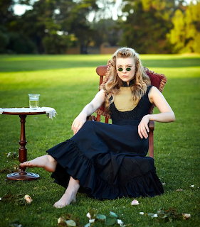 Woman in a black dress sitting barefoot on a chair in a grassy field beside a small table