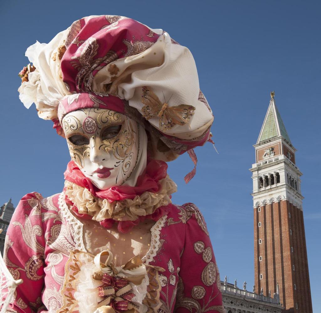 Person in ornate Venice carnival costume with white mask, red gold outfit, and elaborate headpiece against blue sky