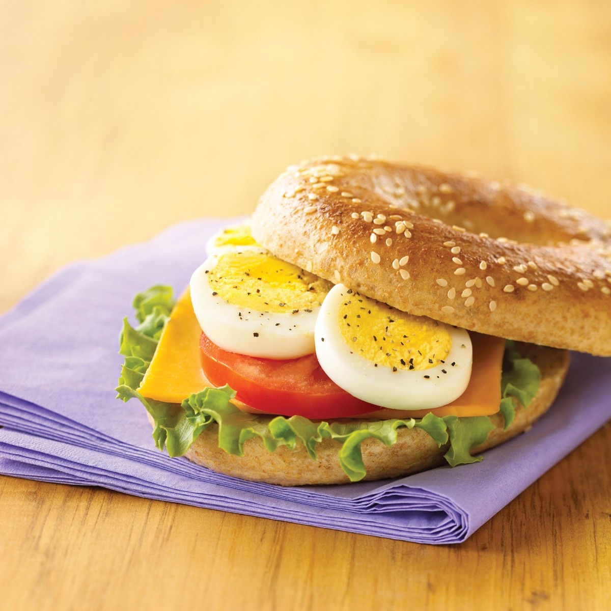 Sesame bagel sandwich with eggs, lettuce, tomato and cheese on purple napkin