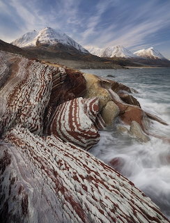 Layered red and white striped rock formations along a rugged arctic coastline with snowy mountains behind