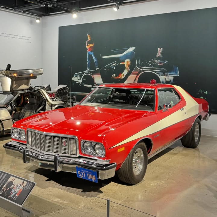 Red vintage Ford Gran Torino and silver DeLorean displayed in museum with dark teal wall and visitors in background