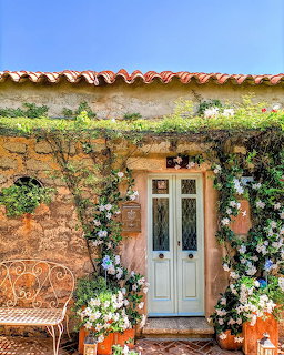 Rustic stone cottage entrance with a white door, vines, flowers, and a bench